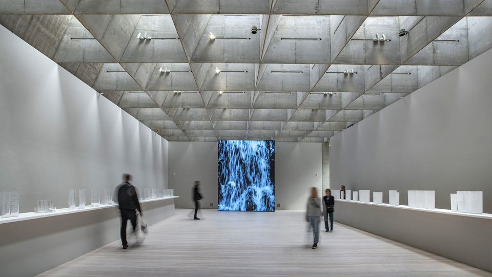 Utställningslokal med ljusinsläpp från taklanterninerna och en digitalskärm med ett konstverk, foto.